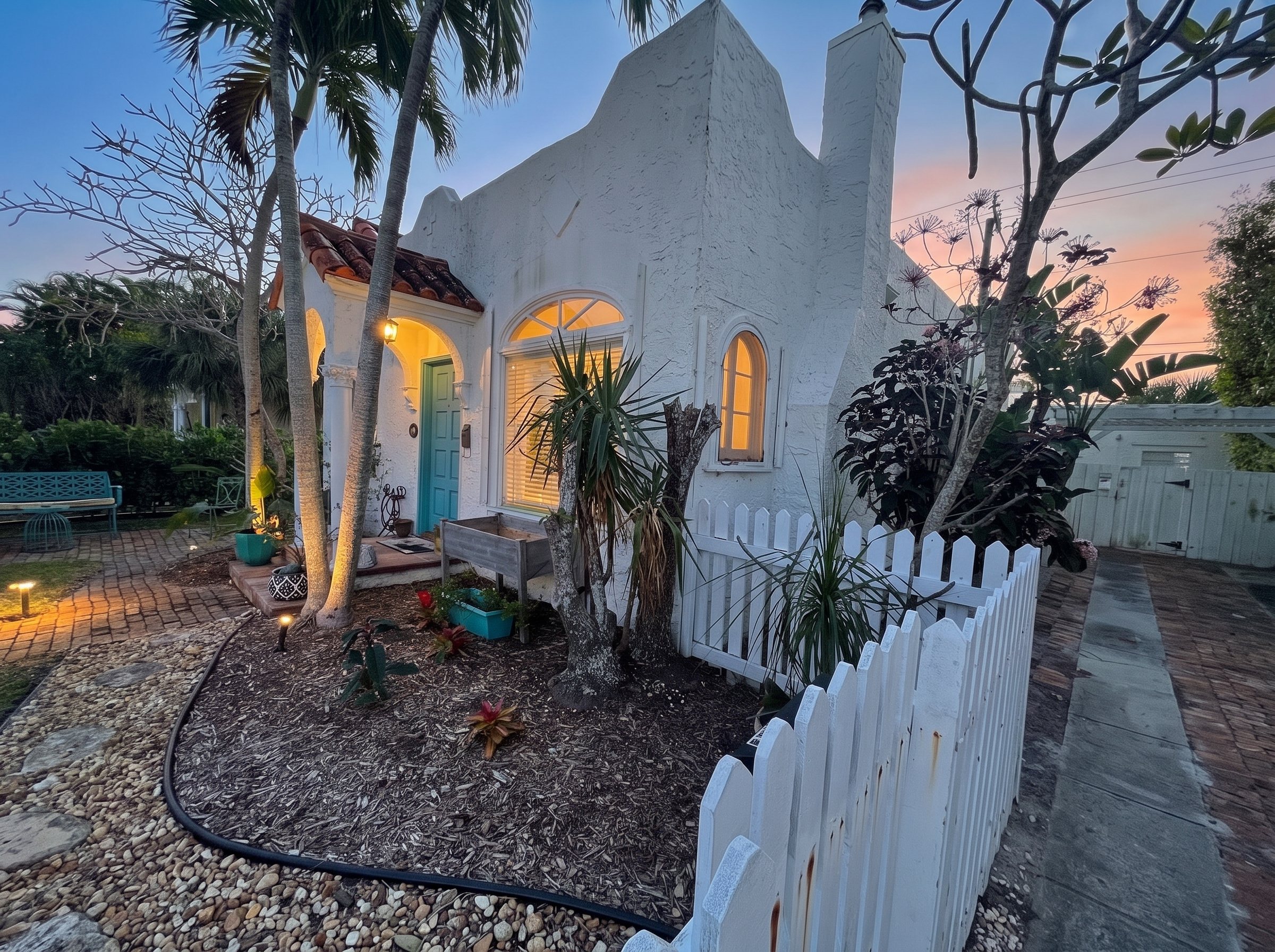 244 Conniston at dusk: white stucco Mediterranean Revival house with warm light in its arched windows, palms and a white picket fence